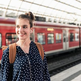 A young woman with a backpack and headphones stands on the platform of a train station.