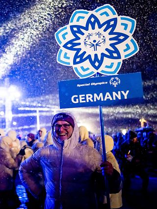 Lächelnder Mann in Winterklamotten hält ein Schild mit dem Logo der Special Olympics Winterspiele und dem Schriftzug ‚Germany‘