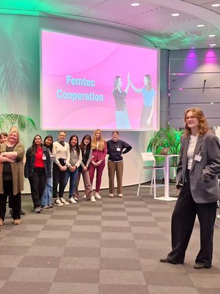 Junge MINT Studentinnen stehen vor einem Präsentations-Bildschirm auf dem „Femtec Cooperation“ steht